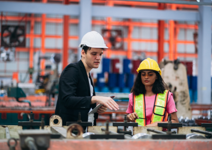 A foreman helps a worker at a metal sheet factory, both in hard hats.