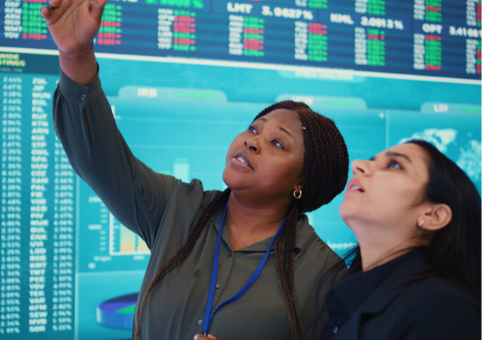 Two people pointing up at something out of shot with a stockmarket live tracker behind them