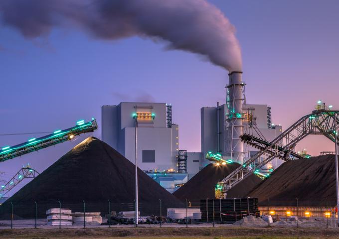 A photo of a coal plant at dusk. Smoke spouts from a chimney in the background while heaps of dirt lie in the foreground.