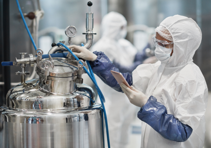 A woman in a hazmat suit stands over a metal container with gloves on, checking the gauge.